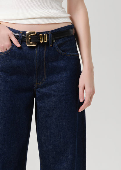 A close-up of a person wearing dark blue denim jeans with a black leather belt featuring a prominent gold buckle, paired with a cream ribbed top against a grey background.