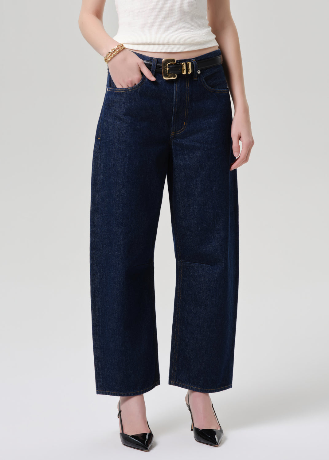 A model wearing dark blue wide-leg cropped jeans with a gold belt, paired with a white top and black pointed-toe heels, shown against a grey background.