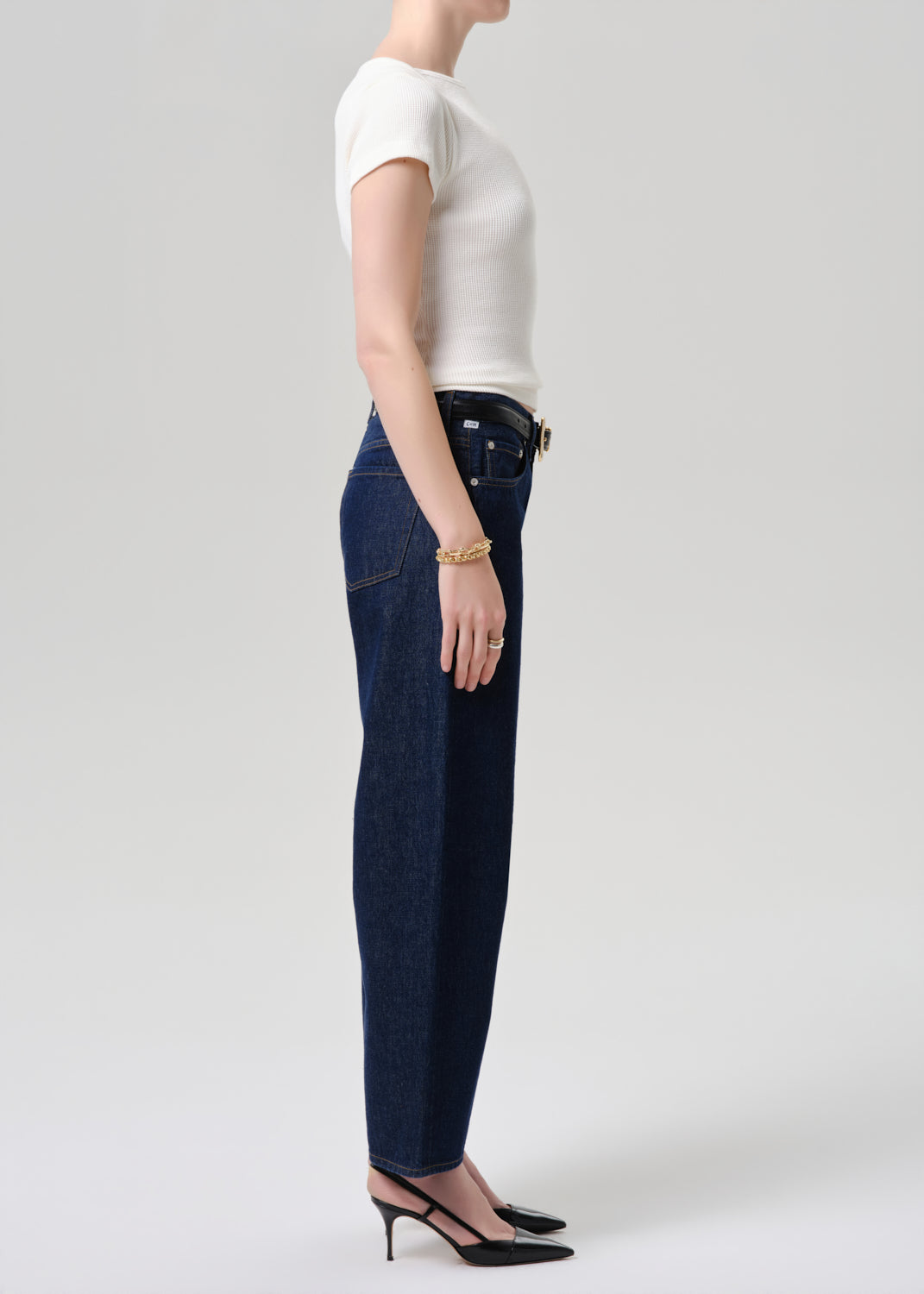 A side-profile studio shot of a model wearing dark blue straight-leg jeans, a white short-sleeve top, and black slingback heels against a grey background.