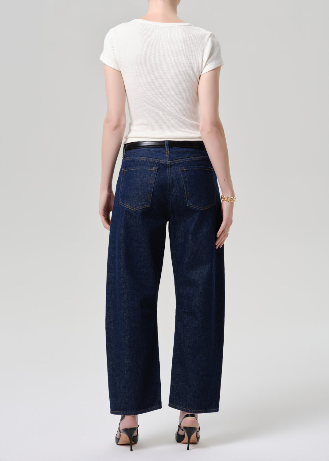 A model shown from the back wearing dark blue wide-leg denim jeans, a white short-sleeve top, and black high heels against a grey background.