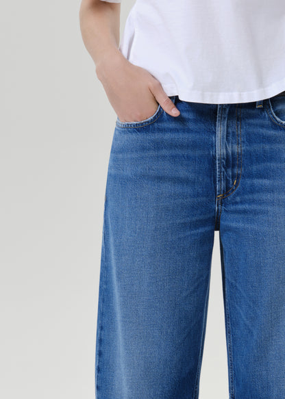 Close-up of a person wearing blue denim jeans and a white top, with one hand in the pocket, against a grey background.