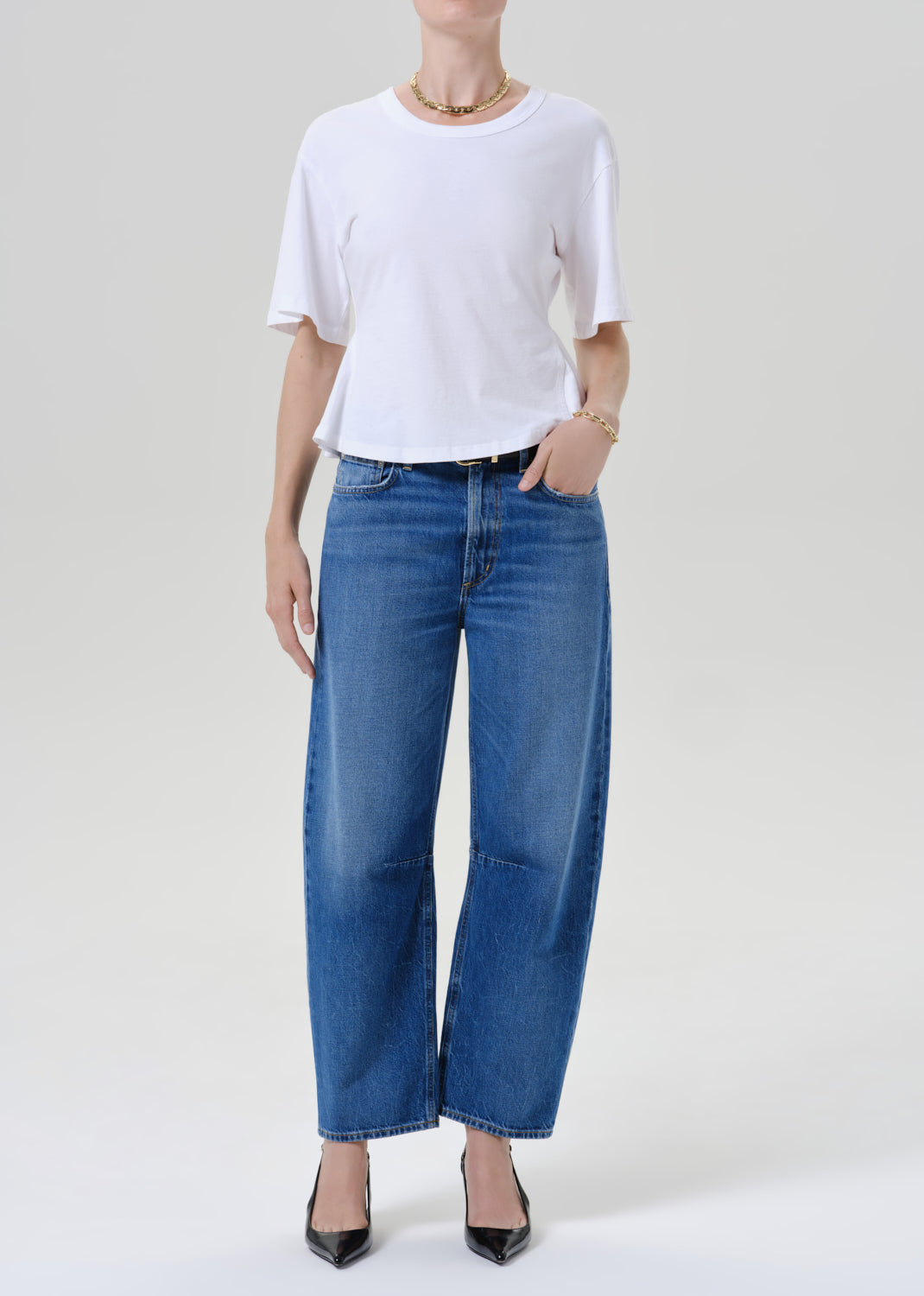 A model wearing a white short-sleeve t-shirt and wide-leg blue denim jeans, paired with black pointed-toe heels, standing against a grey background.