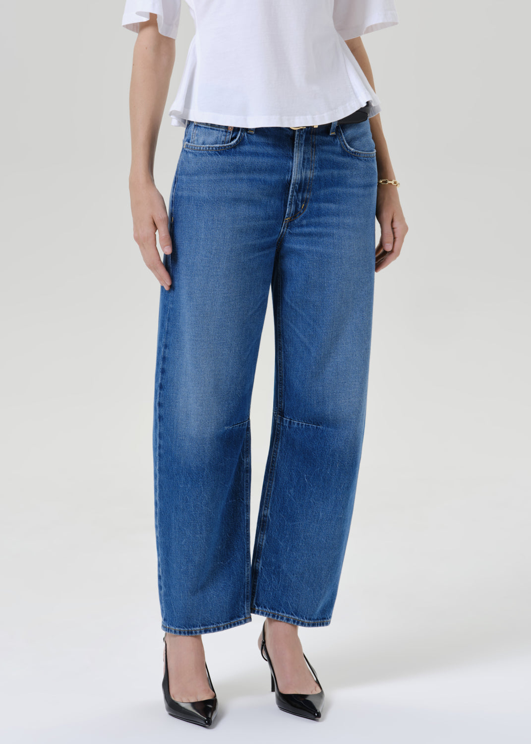 A model wearing a pair of wide-leg cropped blue denim jeans with front patch pockets, styled with a white top and black pointed-toe heels against a grey background.