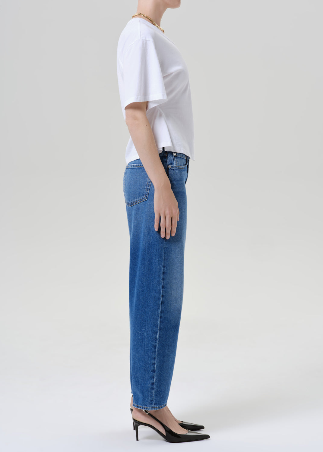 A side-profile view of a model wearing blue straight-leg jeans, a white short-sleeve t-shirt, and black pointed-toe slingback heels against a grey background.