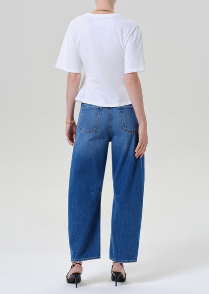 A model shown from the back wearing a white short-sleeve crop top, wide-leg blue denim jeans, and black pointed-toe heels against a grey background.