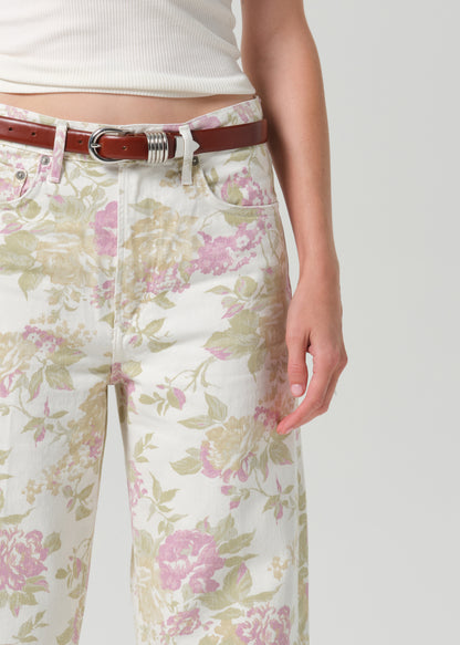 A close-up of a person wearing floral wide-leg jeans with a pink and green print on a cream background, styled with a brown leather belt and a white ribbed top against a grey background.