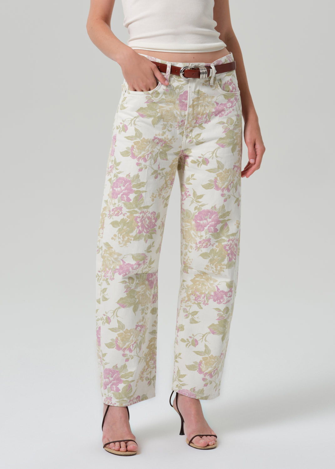 A model wearing a pair of wide-leg cropped jeans with a pink and green floral pattern on a cream background, styled with a brown belt, white crop top, and black strappy heels against a grey background.