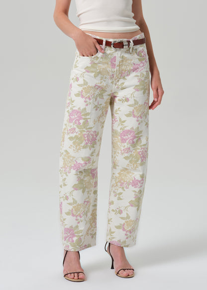 A model wearing a pair of wide-leg cropped jeans with a pink and green floral pattern on a cream background, styled with a brown belt, white crop top, and black strappy heels against a grey background.