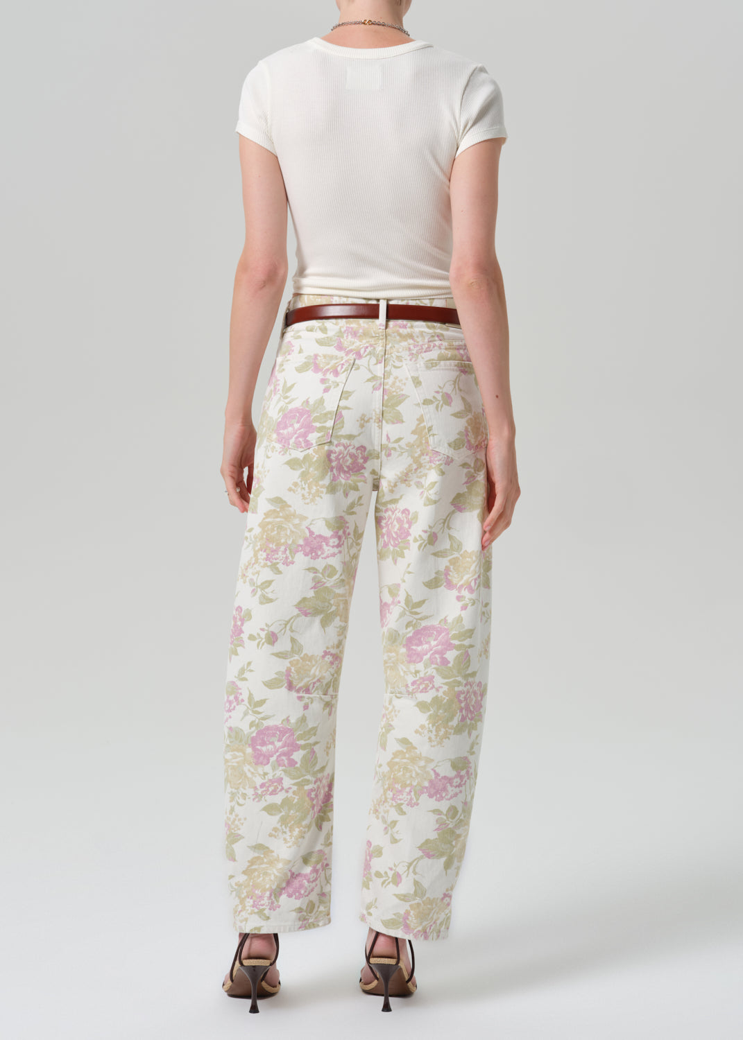 A model shown from the back wearing cream wide-leg trousers with a pink and green floral pattern, styled with a brown belt, a white t-shirt, and brown heels against a grey background.