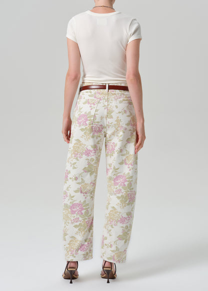 A model shown from the back wearing cream wide-leg trousers with a pink and green floral pattern, styled with a brown belt, a white t-shirt, and brown heels against a grey background.