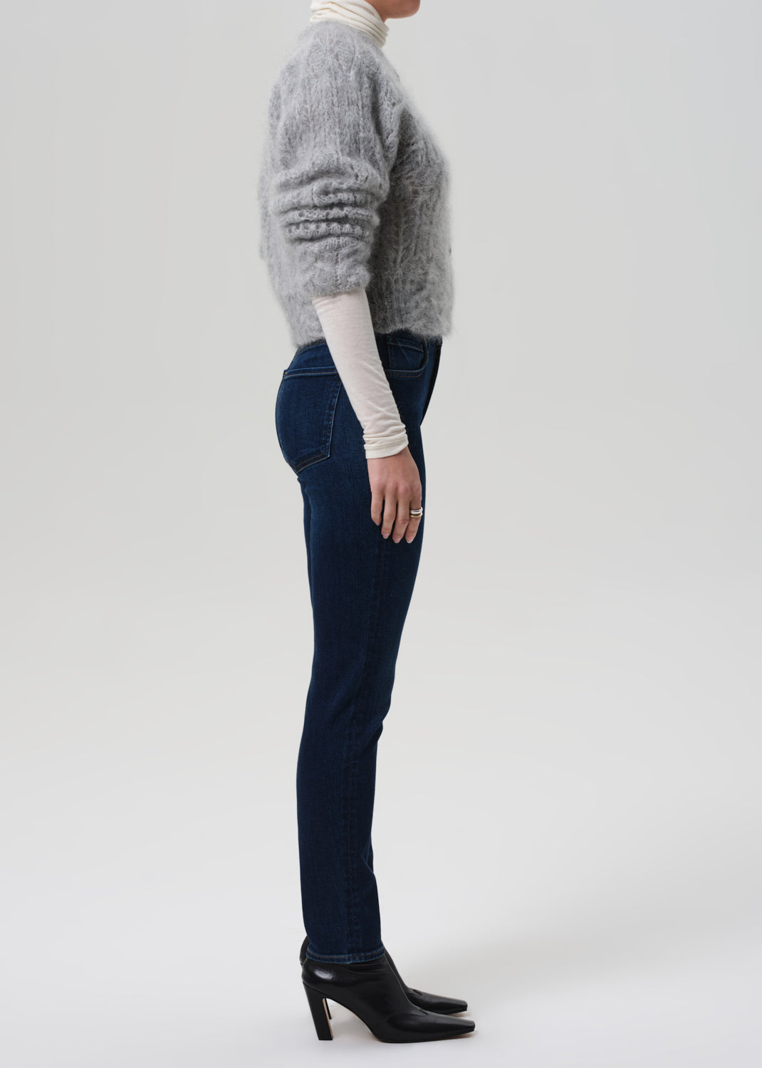 Person wearing a gray sweater, white turtleneck, dark jeans, and black shoes on a light gray background