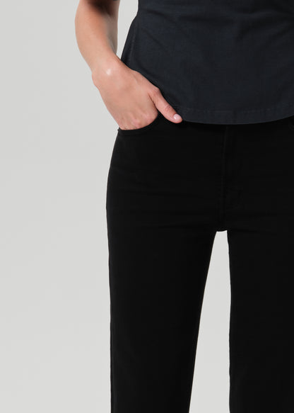 Close-up of a person wearing black pants and a black top, with one hand in the pocket, against a grey background.