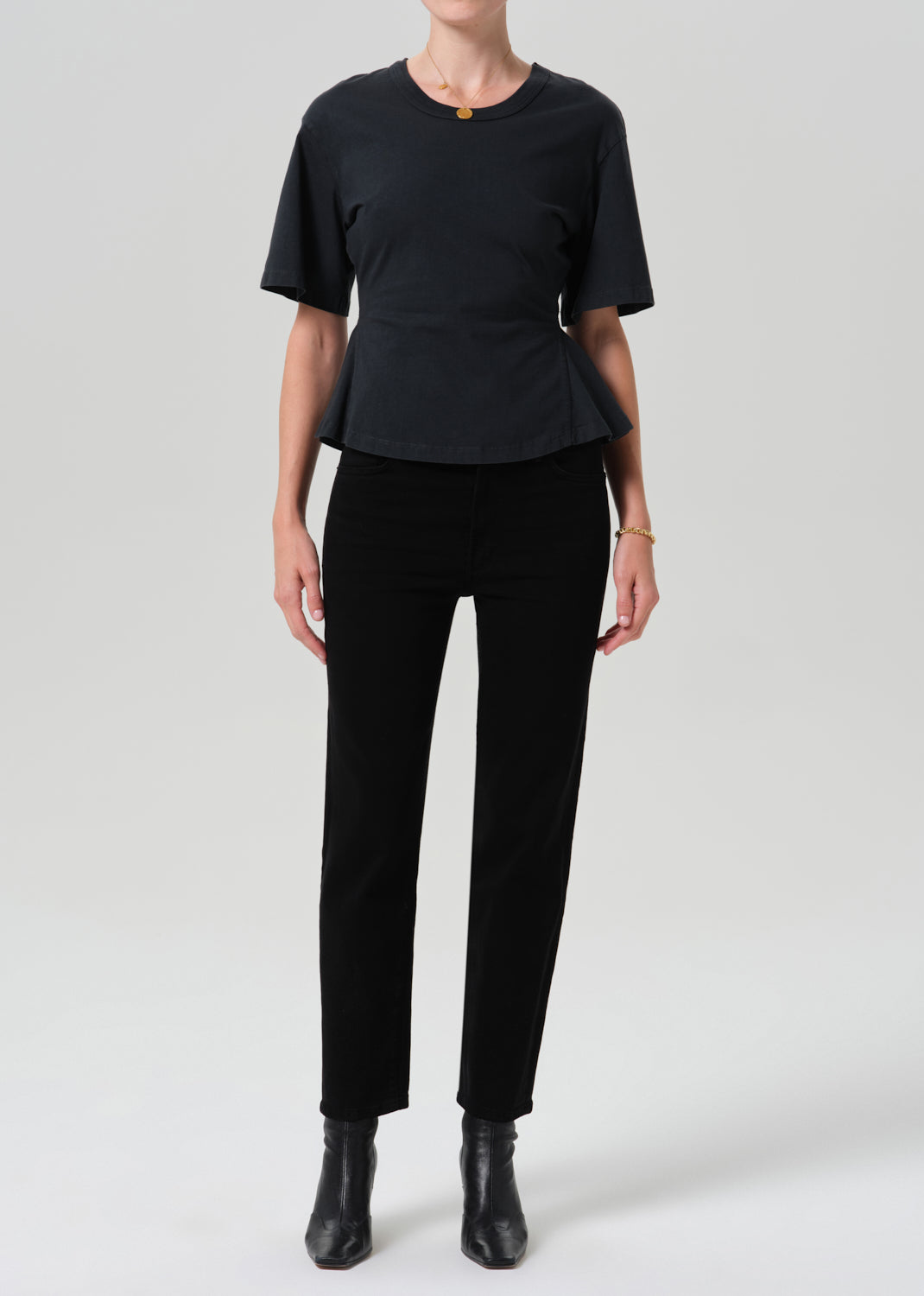 A model wearing a black short-sleeve peplum top, black trousers, and black ankle boots, standing against a grey background.