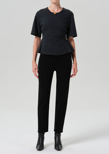 A model wearing a black short-sleeve peplum top, black trousers, and black ankle boots, standing against a grey background.