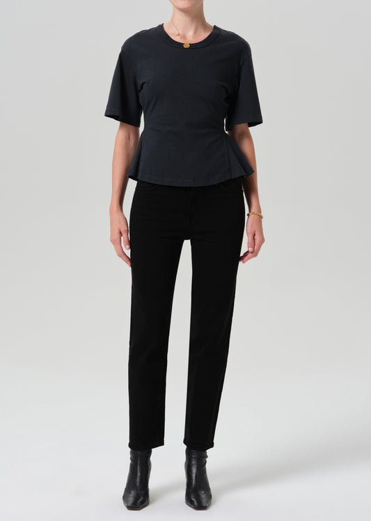 A model wearing a black short-sleeve peplum top, black trousers, and black ankle boots, standing against a grey background.