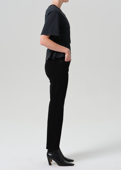A model shown in profile wearing a black short-sleeve top, black slim-fit trousers, and black heeled ankle boots against a grey background.