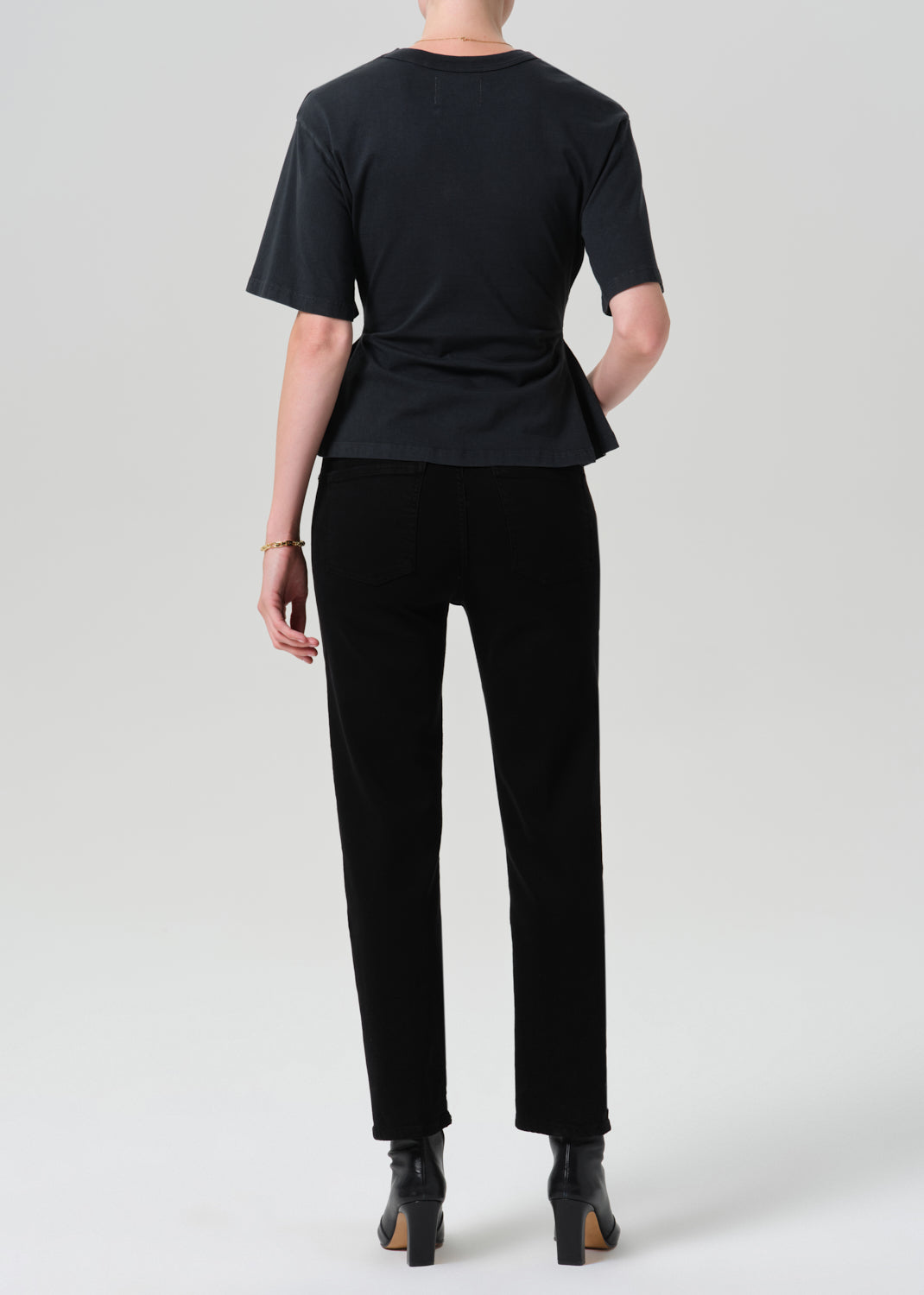 A model shown from the back wearing a black short-sleeve t-shirt with a peplum waist, black trousers, and black ankle boots, photographed against a grey background.