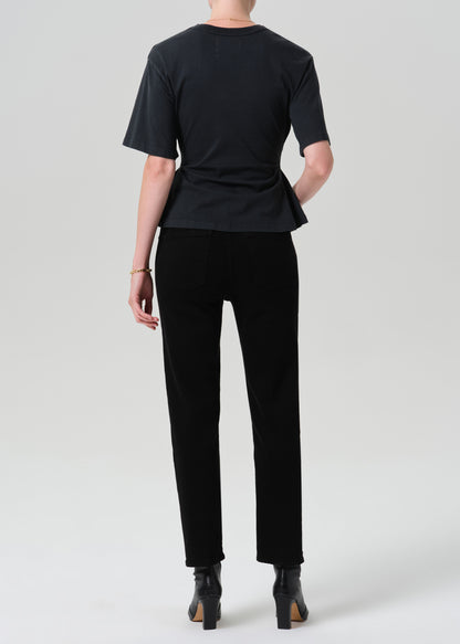 A model shown from the back wearing a black short-sleeve t-shirt with a peplum waist, black trousers, and black ankle boots, photographed against a grey background.