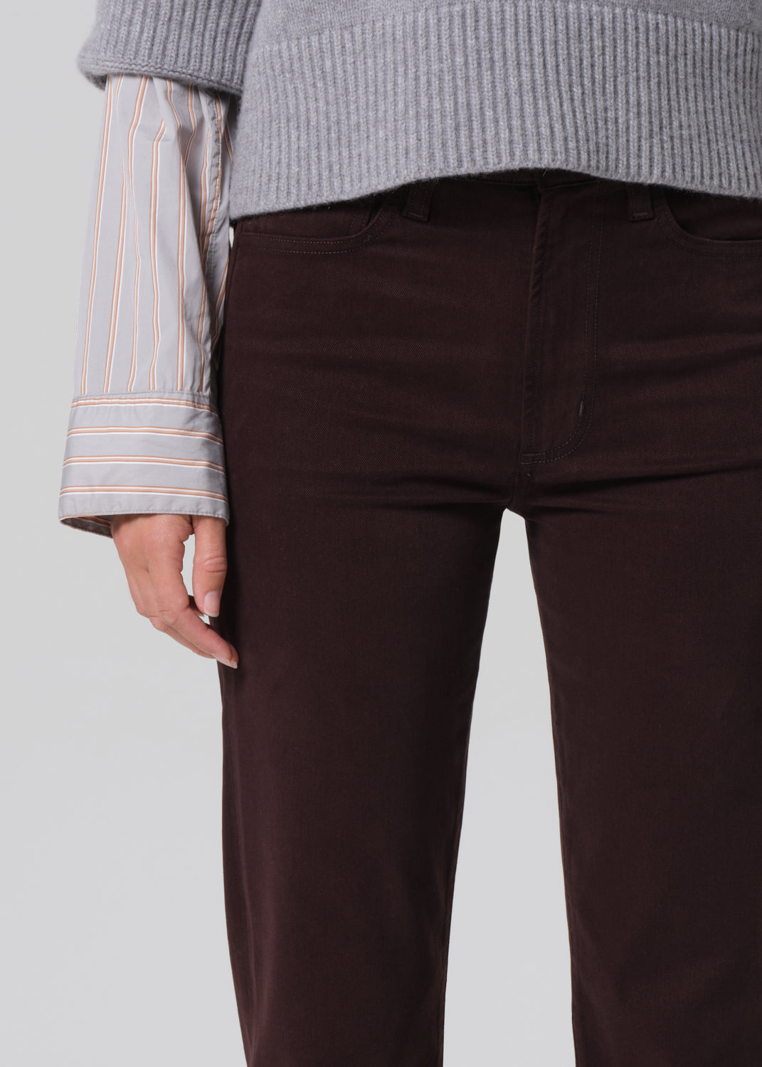 Brown pants worn with a gray sweater and striped shirt on a plain background