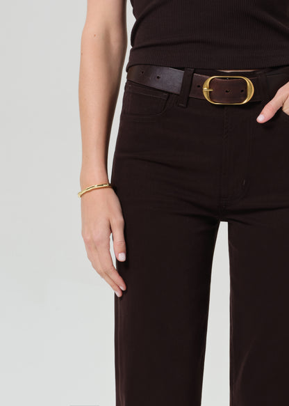 Person wearing a brown sleeveless top and brown pants with a belt on a grey background