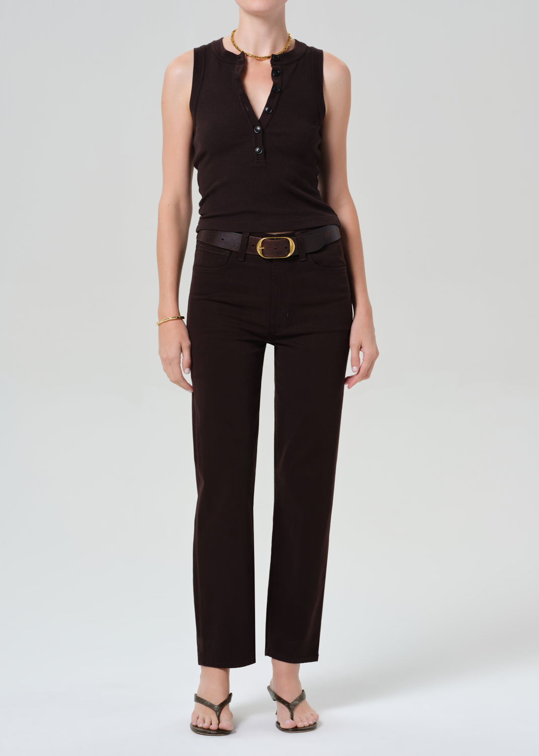 Person wearing a brown sleeveless top and brown pants with a belt on a grey background