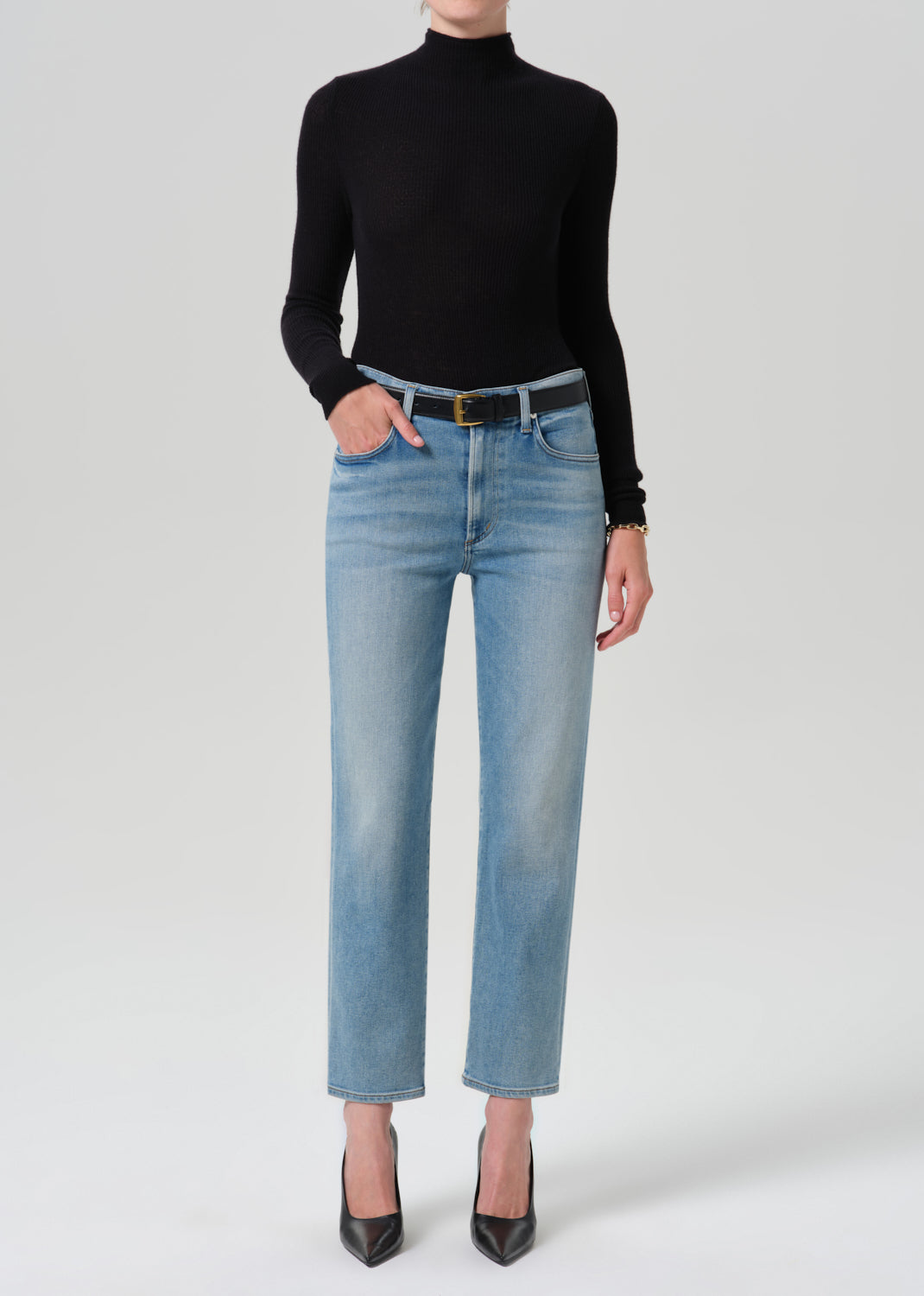 A model wearing a black long-sleeve mock neck top, light-wash blue straight-leg jeans with a black belt, and black pointed-toe heels, standing against a grey background.