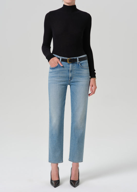 A model wearing a black long-sleeve mock neck top, light-wash blue straight-leg jeans with a black belt, and black pointed-toe heels, standing against a grey background.