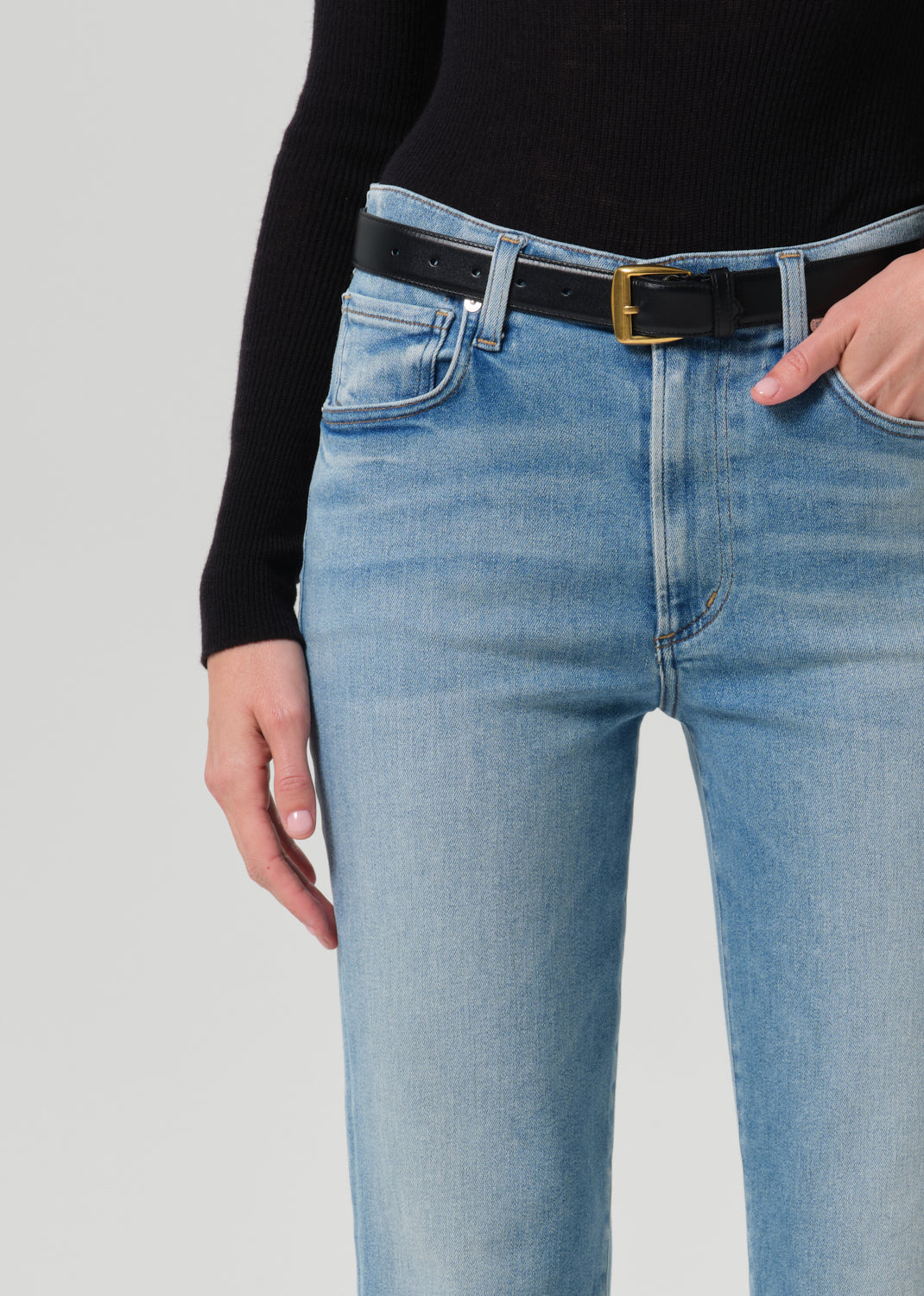Close-up of a person wearing light-wash blue jeans with a black belt featuring a gold buckle, paired with a black ribbed long-sleeve top against a grey background.