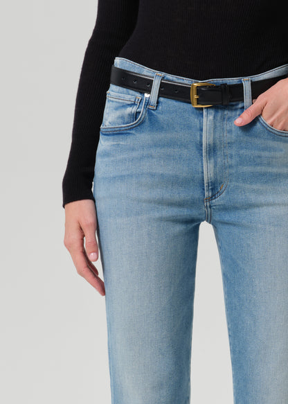 Close-up of a person wearing light-wash blue jeans with a black belt featuring a gold buckle, paired with a black ribbed long-sleeve top against a grey background.