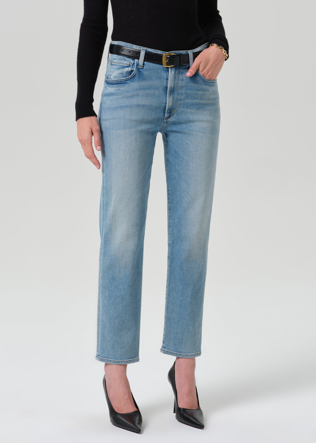 A model wearing light-wash blue cropped jeans with a black belt, paired with a black long-sleeve top and black pointed-toe heels, photographed against a grey background.