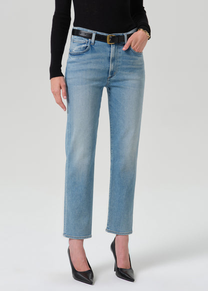 A model wearing light-wash blue cropped jeans with a black belt, paired with a black long-sleeve top and black pointed-toe heels, photographed against a grey background.