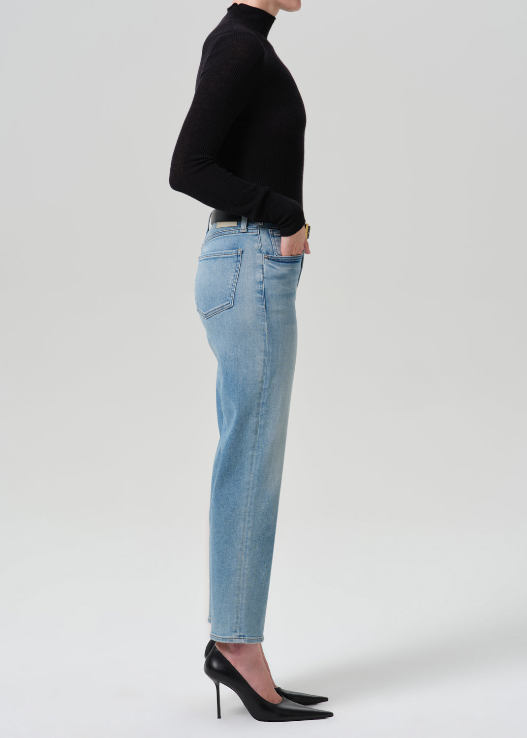 A side-profile studio shot of a model wearing light-wash blue jeans, a black long-sleeve mock-neck top, and black pointed-toe heels against a grey background.