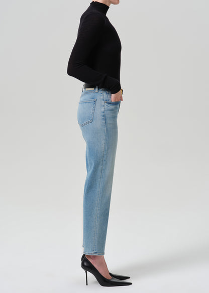 A side-profile studio shot of a model wearing light-wash blue jeans, a black long-sleeve mock-neck top, and black pointed-toe heels against a grey background.