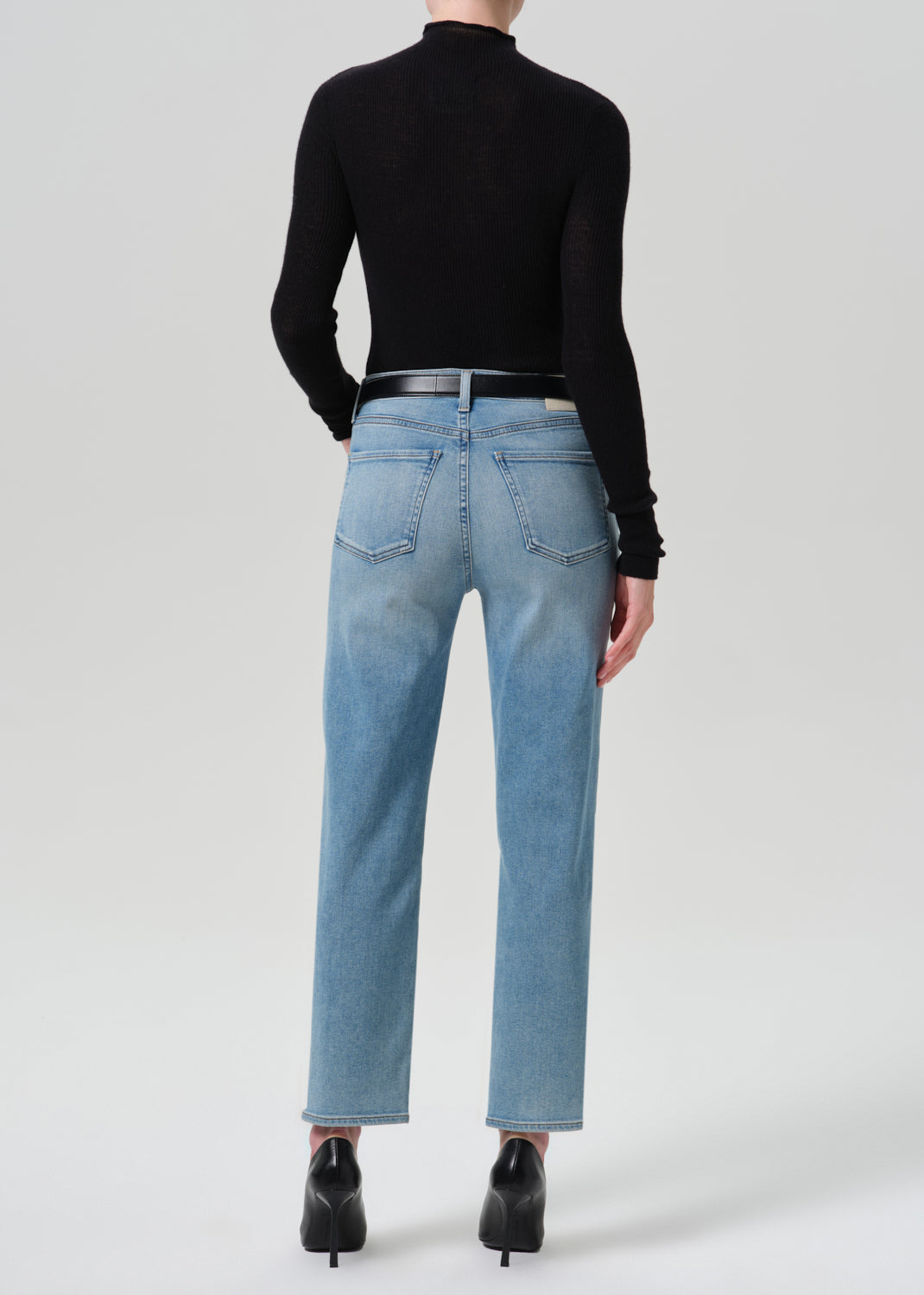 A model shown from the back wearing light-wash blue straight-leg jeans, a black long-sleeve mock-neck top, a black belt, and black pointed-toe heels against a grey background.
