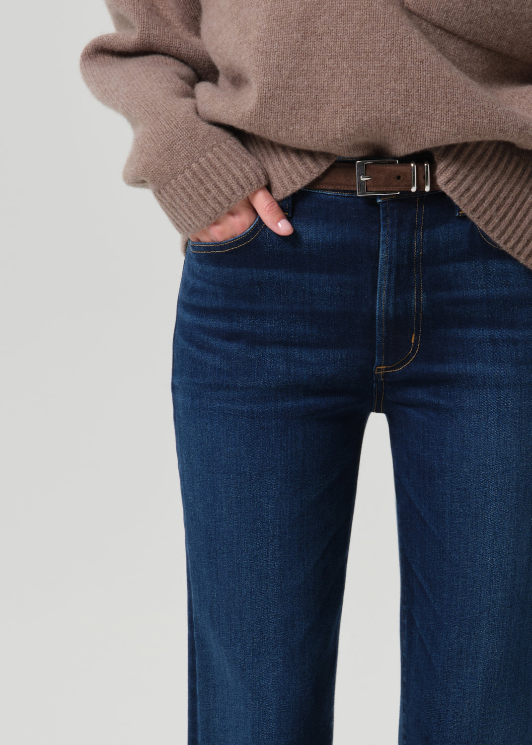Close-up of a person wearing dark blue denim jeans with a brown belt, paired with a taupe knit sweater, against a grey background.