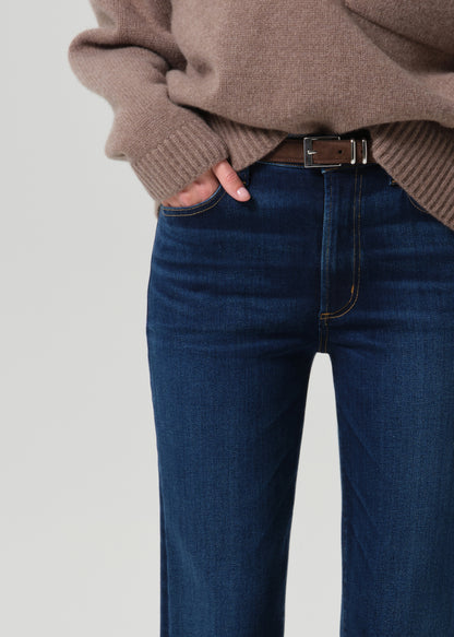 Close-up of a person wearing dark blue denim jeans with a brown belt, paired with a taupe knit sweater, against a grey background.