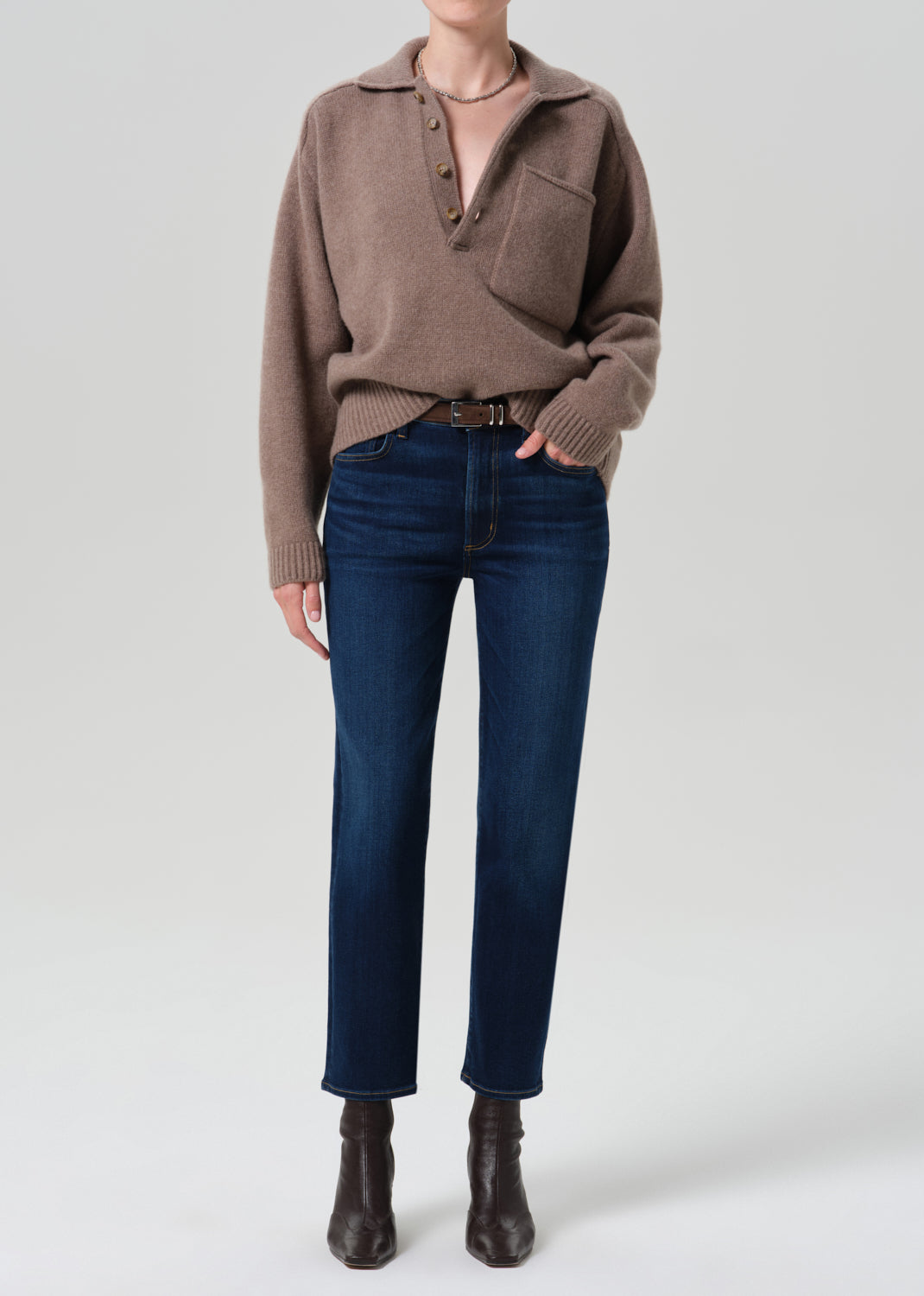 A model wearing a taupe brown knit polo sweater, dark blue straight-leg jeans, and dark brown leather ankle boots, standing against a grey background.