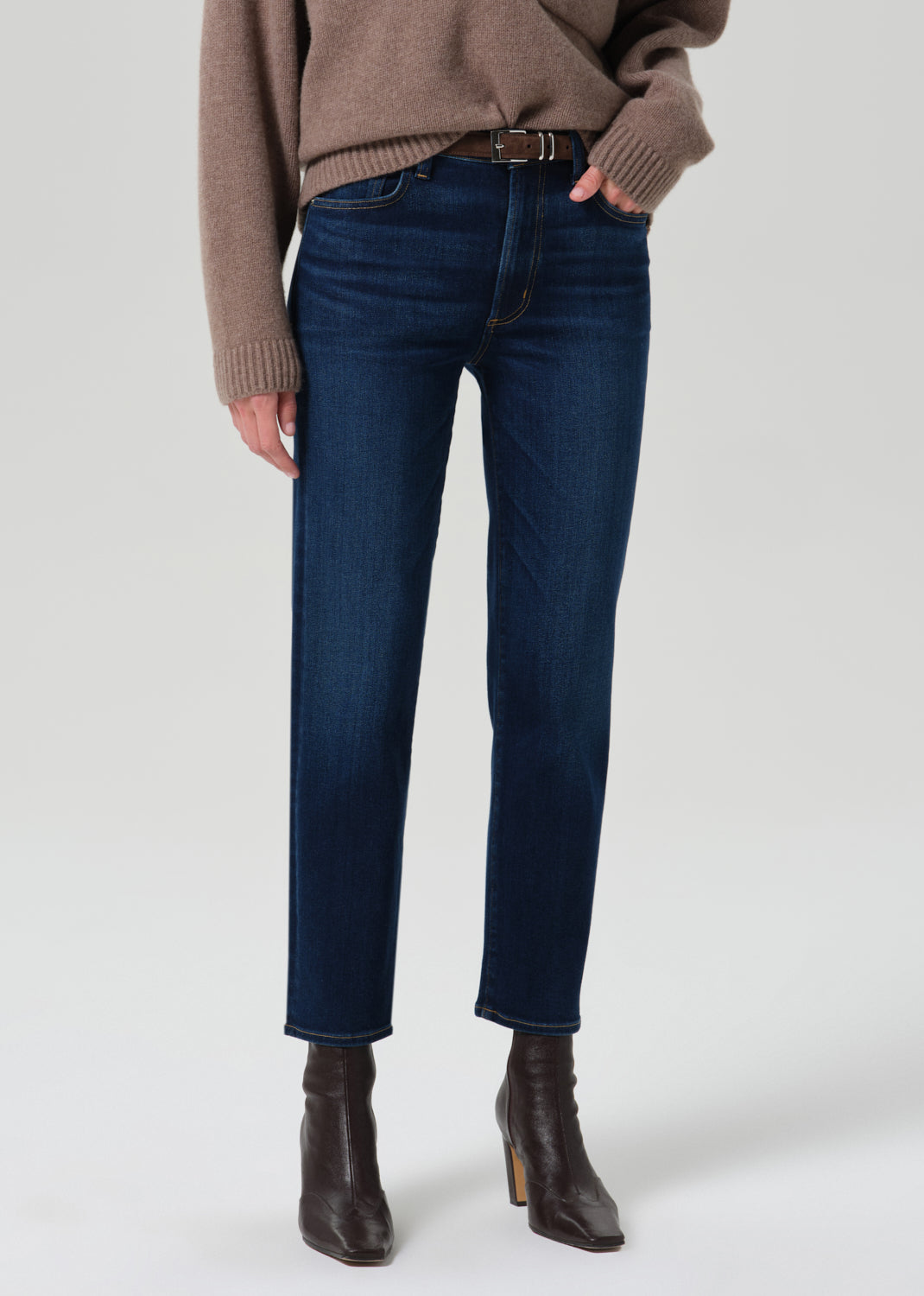 A model wearing dark blue straight-leg jeans, styled with a brown knit sweater and dark brown heeled ankle boots, shown from the waist down on a grey background.