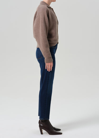 A side-profile studio shot of a model wearing a taupe knit polo cardigan, dark blue jeans, and dark brown leather heeled ankle boots against a grey background.