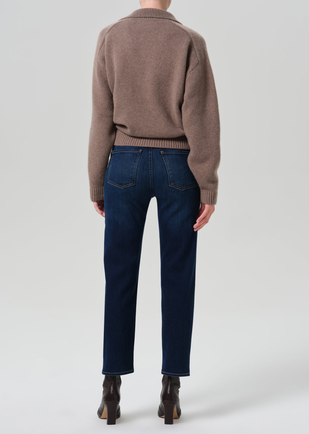 Rear view of a model wearing a taupe knit sweater and dark blue straight-leg jeans with brown ankle boots, shot against a grey background.