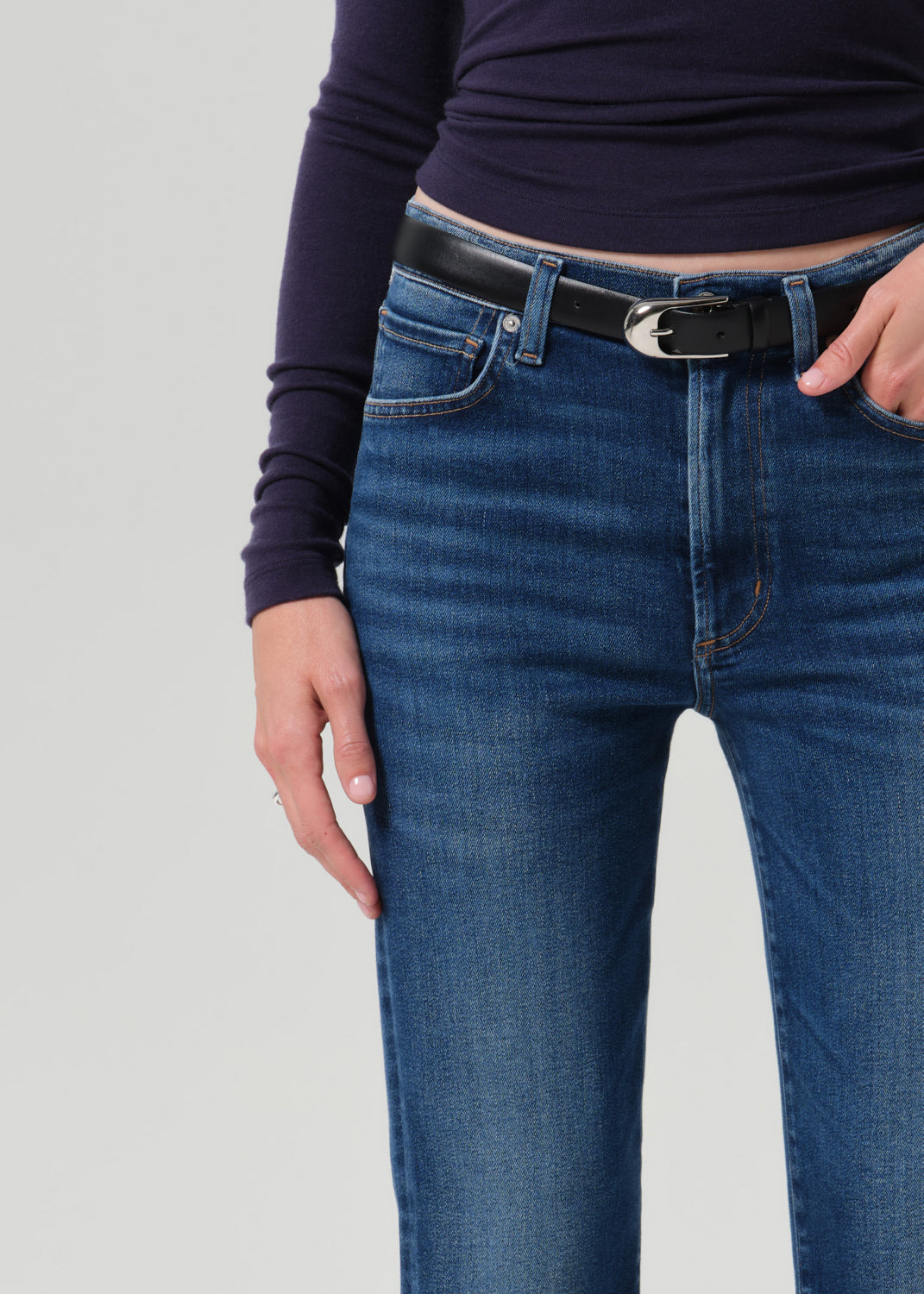 A close-up of a person wearing blue denim jeans with a black belt and a dark purple long-sleeve top, shown from the waist down against a grey background.