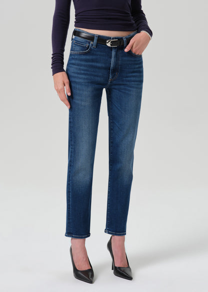 A model wearing a pair of dark blue straight-leg jeans with a black belt, paired with a dark long-sleeve top and black heels, shown against a grey background.