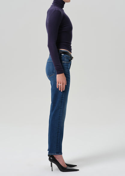 A side-profile studio shot of a model wearing dark blue straight-leg jeans, a dark purple long-sleeve mock neck top, and black pointed-toe heels against a grey background.