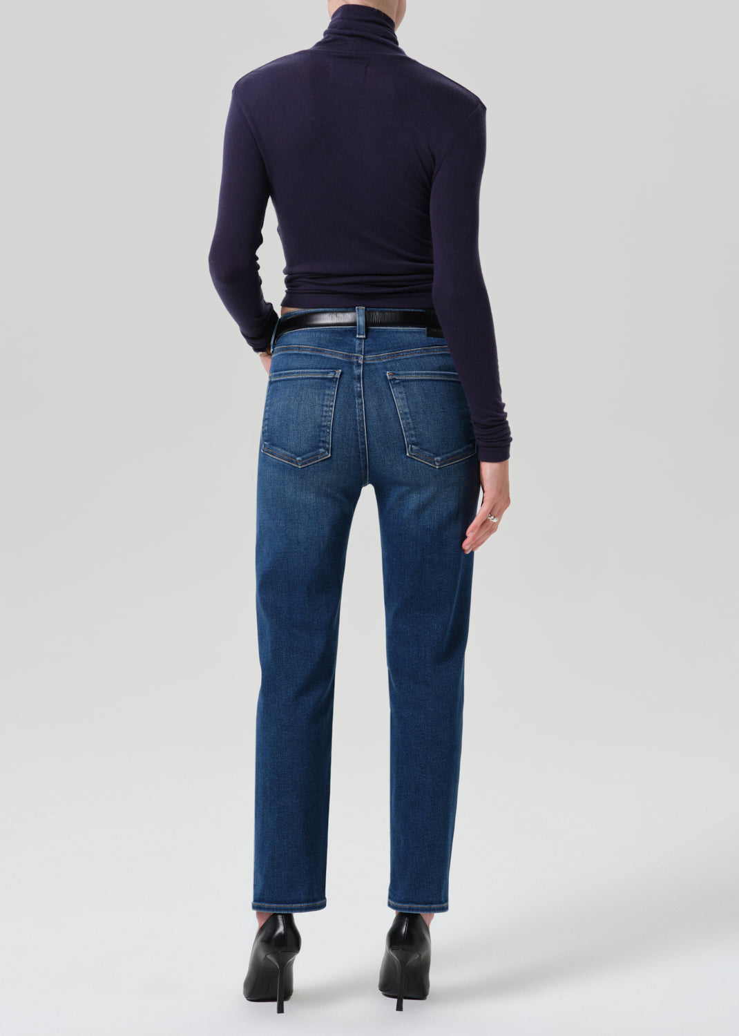 A model shown from the back wearing dark blue straight-leg jeans, a dark navy turtleneck top, a black belt, and black high-heeled boots against a grey background.