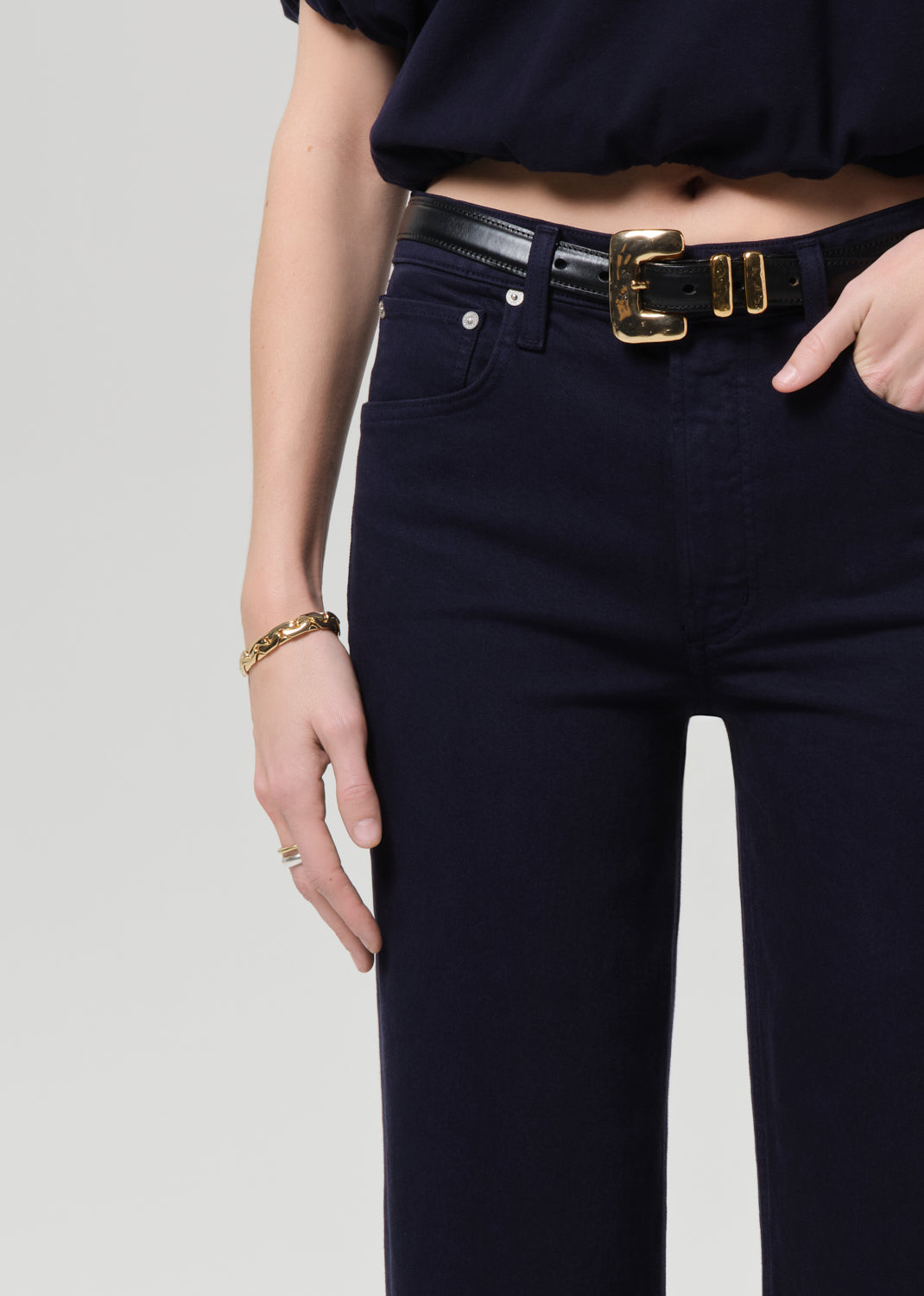 Person wearing navy blue jeans with a gold belt buckle on a light gray background