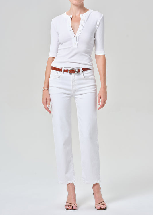 A model wearing a white short-sleeve henley top and white cropped jeans with a brown leather belt, paired with brown heeled sandals, against a grey background.