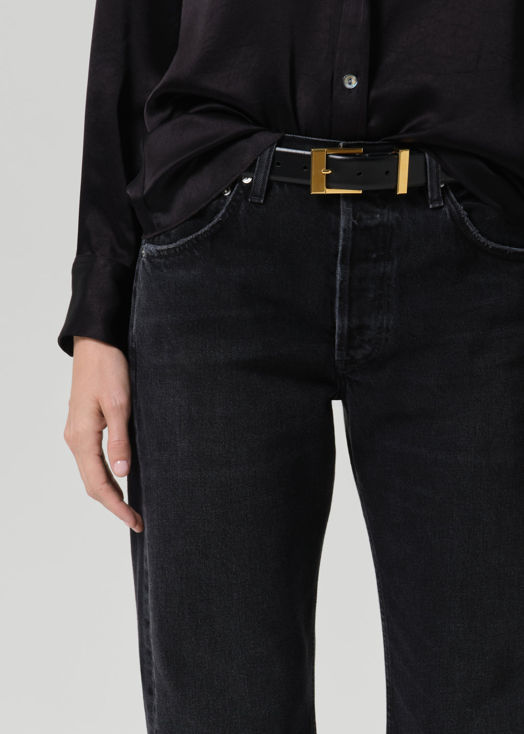 Close-up of a person wearing black jeans with a black belt featuring a gold rectangular buckle, paired with a black shirt against a grey background.