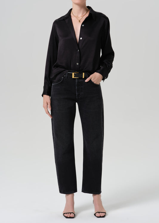 A model wearing a black satin long-sleeve button-up shirt, black wide-leg jeans with a gold belt, and black heeled sandals, photographed against a grey background.