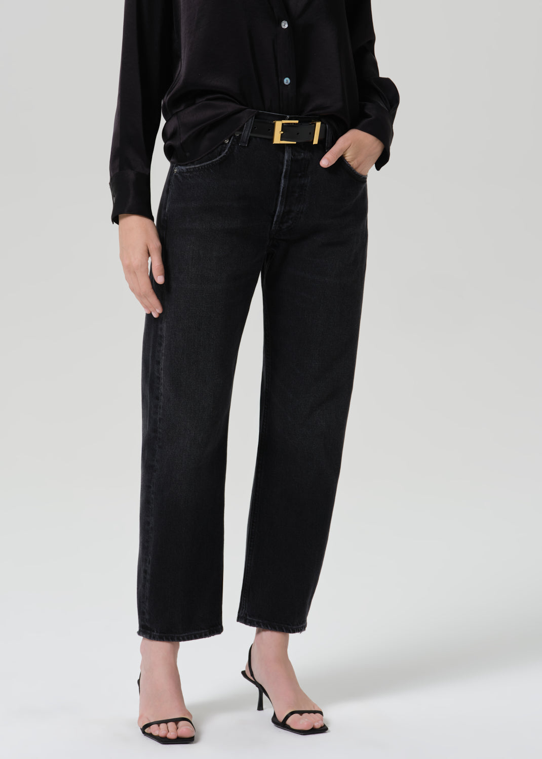 A model wearing black wide-leg cropped jeans, a black long-sleeve button-up shirt, a black belt with a gold buckle, and black strappy high-heeled sandals, photographed against a grey background.