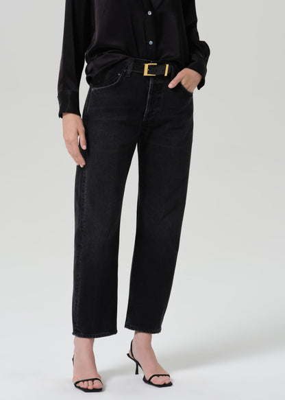 A model wearing black wide-leg cropped jeans, a black long-sleeve button-up shirt, a black belt with a gold buckle, and black strappy high-heeled sandals, photographed against a grey background.